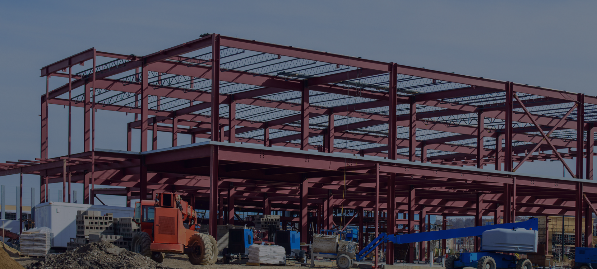 Red steel-frame building under construction with trucks and workers beneath a partly roofed structure
