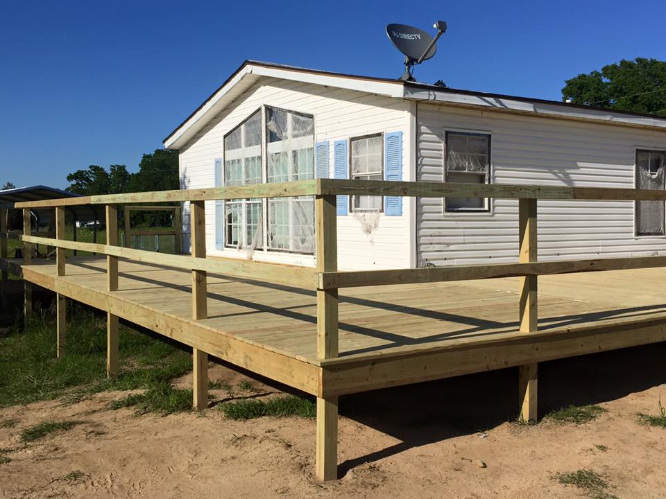 White house with a large wooden deck and railing on a sandy yard, with a satellite dish on the roof