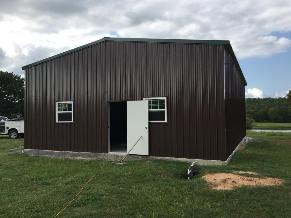 Brown metal barn with white door and windows in a grassy field under a cloudy sky