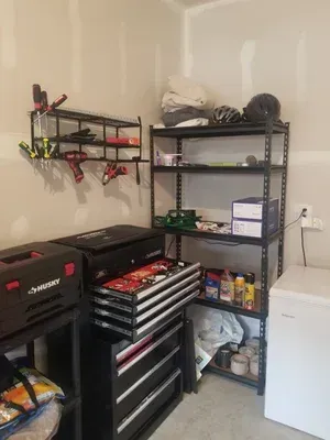 Garage workshop with black tool chest, shelving, wall-mounted tools, and a white refrigerator or freezer