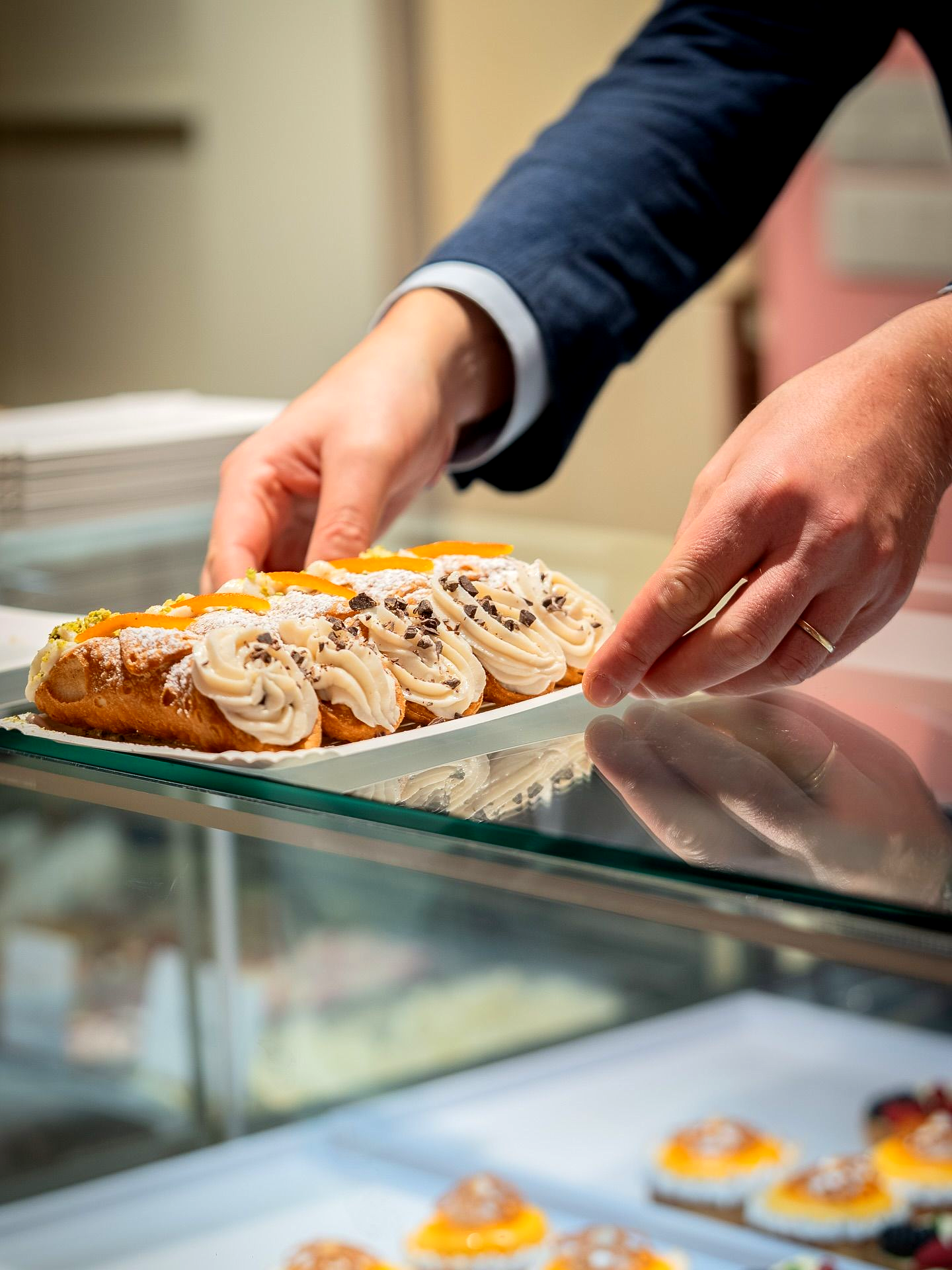 Hands placing a pastry, topped with cream and fruit, on a shelf inside a glass display case.Domenica 
Home made italian pastries, food & coffee 
