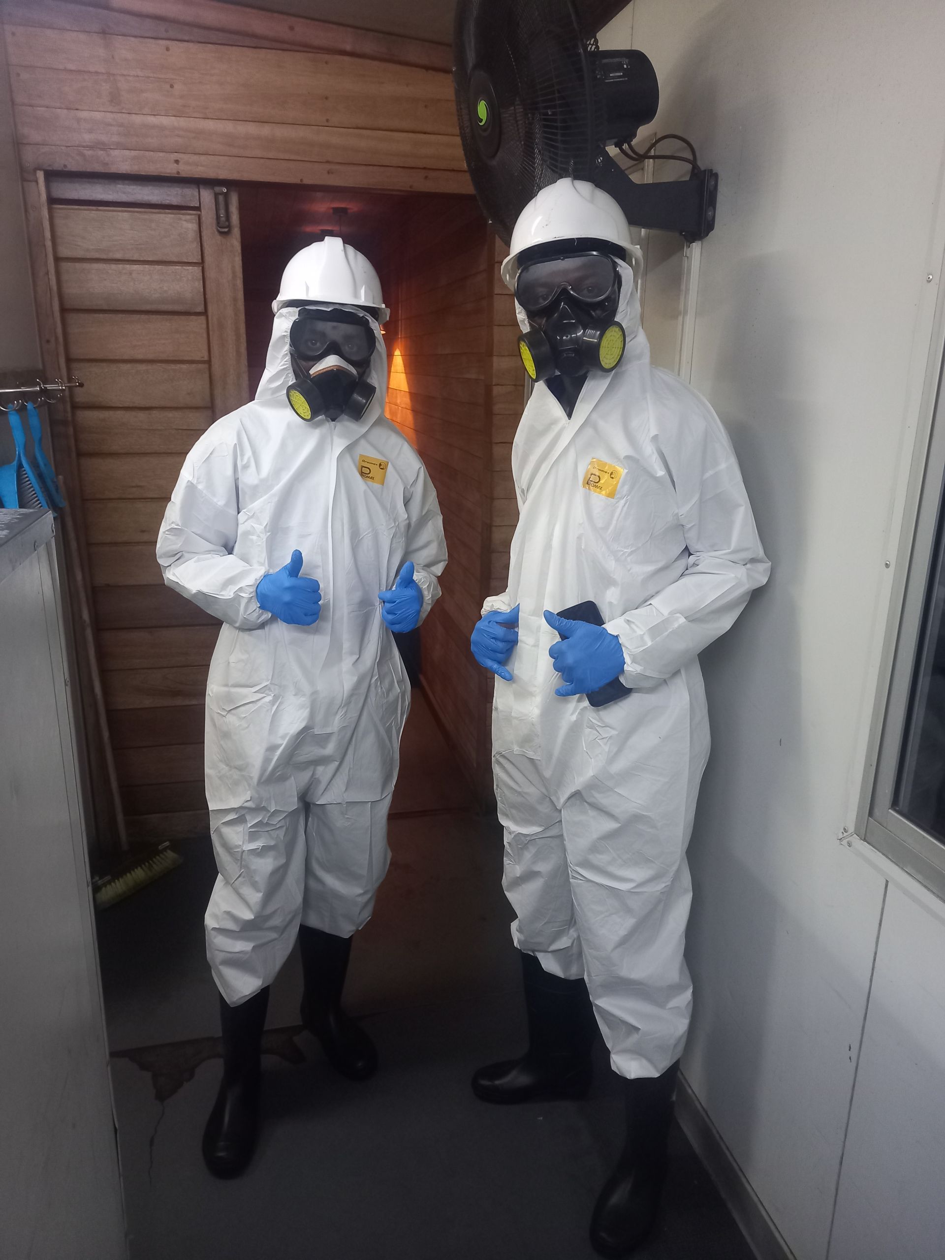 Two people in white hazmat suits, thumbs up, wearing respirators and goggles in a room.