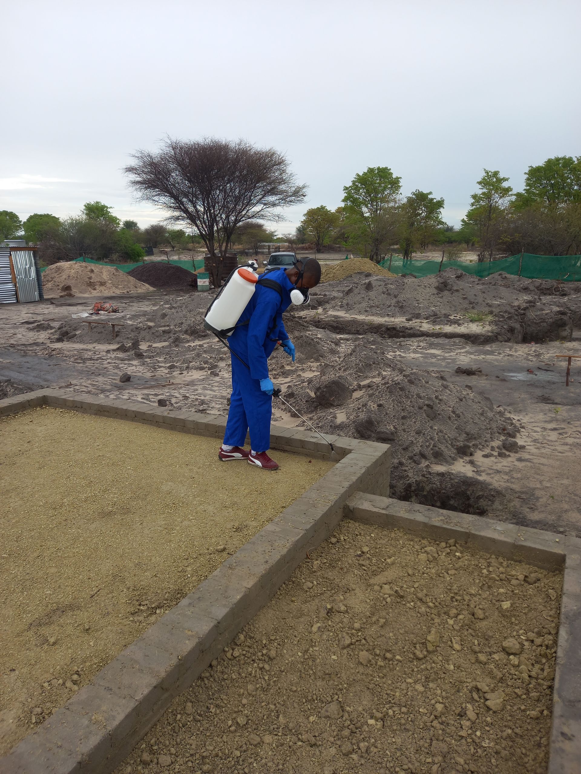 Person in blue suit sprays area with a tank sprayer, outdoors, gravel-filled concrete structures.