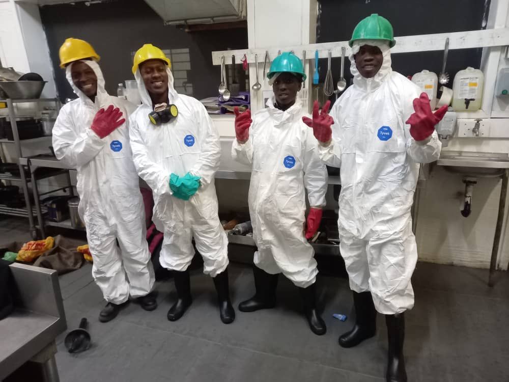 Four people in white hazmat suits, boots, and hard hats, posing in a kitchen, wearing gloves.