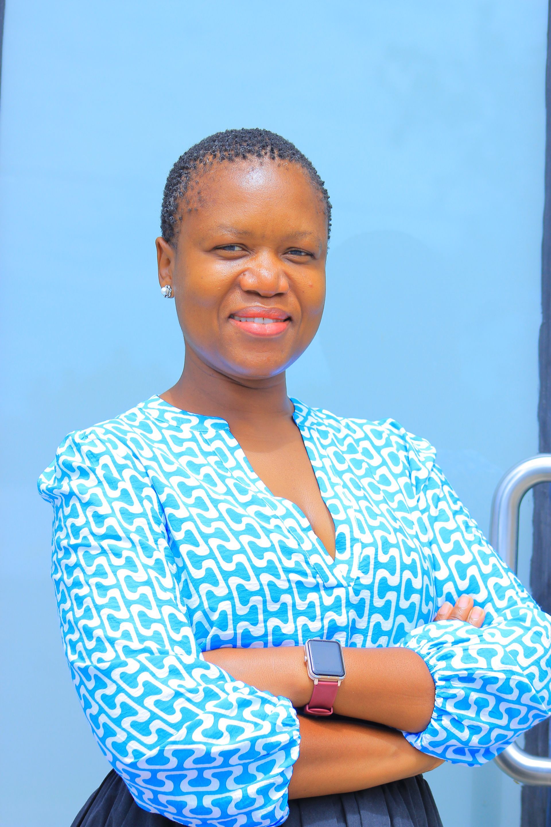 Woman in blue patterned blouse with arms crossed, smiling, in front of a blue wall and a door.