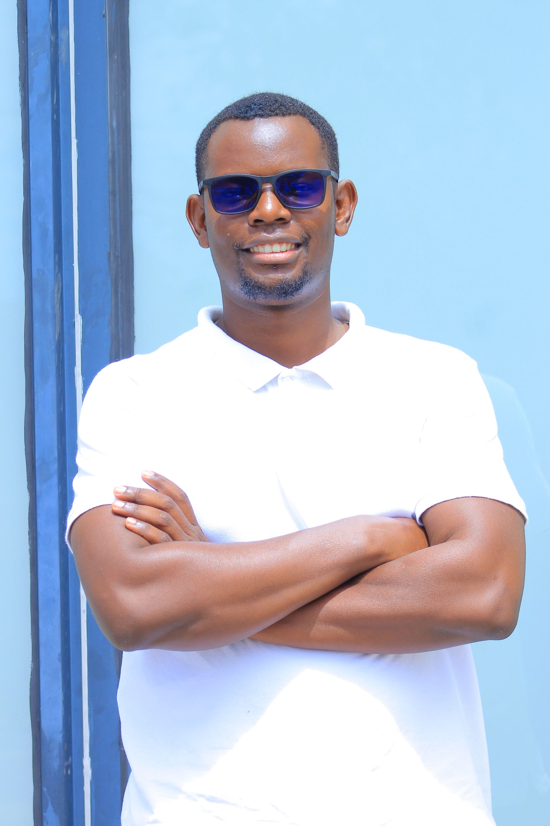 Man in sunglasses and white shirt, arms crossed, stands in front of a blue background.