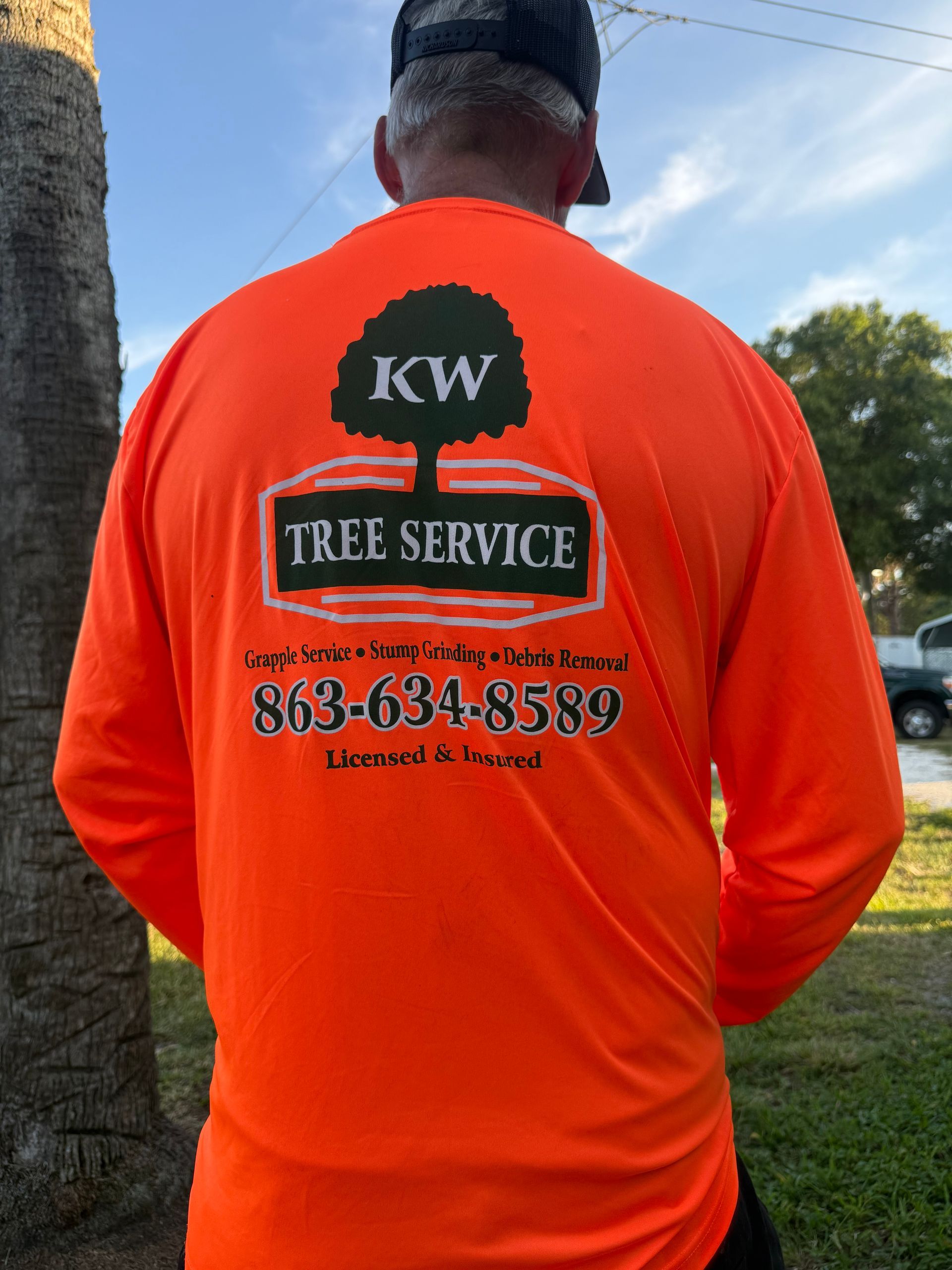 Person in an orange KW Tree Service shirt standing outdoors near a tree.