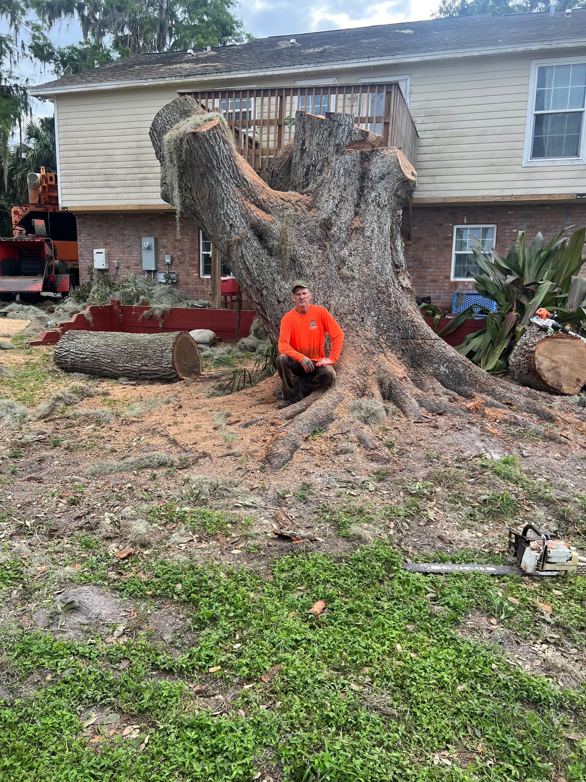 Person in orange jacket sitting beside a large uprooted tree in a front yard after storm damage
