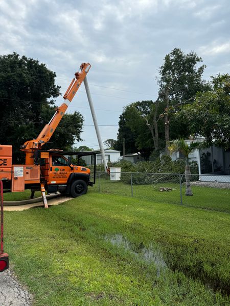 Orange crane truck lifting a long white pole in a grassy yard with trees and a house in the background Orange crane truck lifting a long white pole in a grassy yard with trees and a house in the background