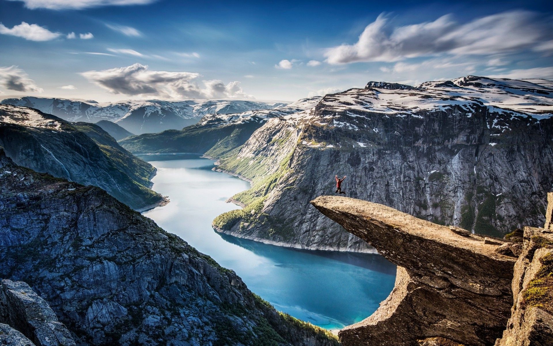 Person standing on a cliff over a blue fjord between steep mountains under a cloudy sky