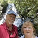 Two smiling people in red shirts posing in front of a waterfall in a lush forest