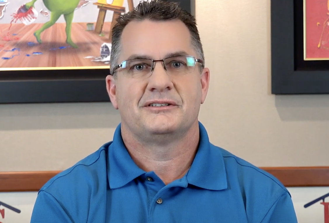 Man with glasses in a blue shirt, centered in front of a wall with colorful paintings.