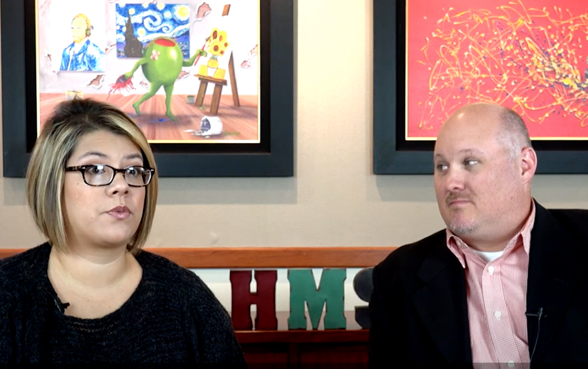 Woman and man seated, speaking, in front of paintings and decorative letters.