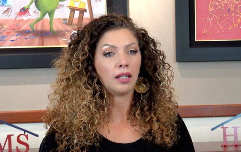 Woman with curly brown hair speaks, wearing black top, in front of artwork and business logo.
