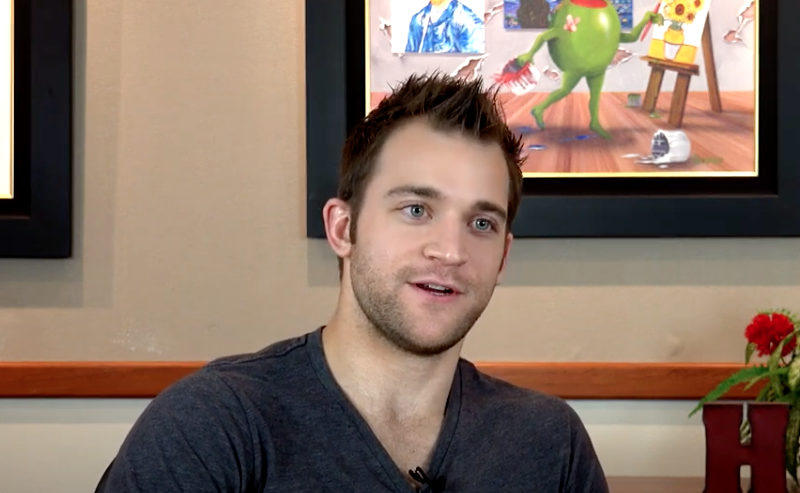 A man with short brown hair and a gray shirt smiles while speaking. A painting of a frog is behind him.