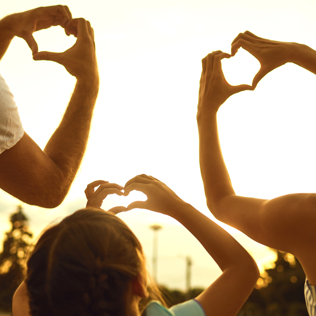 Three people making heart shapes with their hands against a sunset.