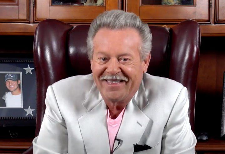 Man with gray hair and mustache smiles, wearing a light suit and pink shirt, seated in a leather chair.