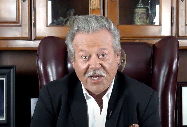 Man in a suit, seated in a leather chair, speaking. Behind him are wooden cabinets.