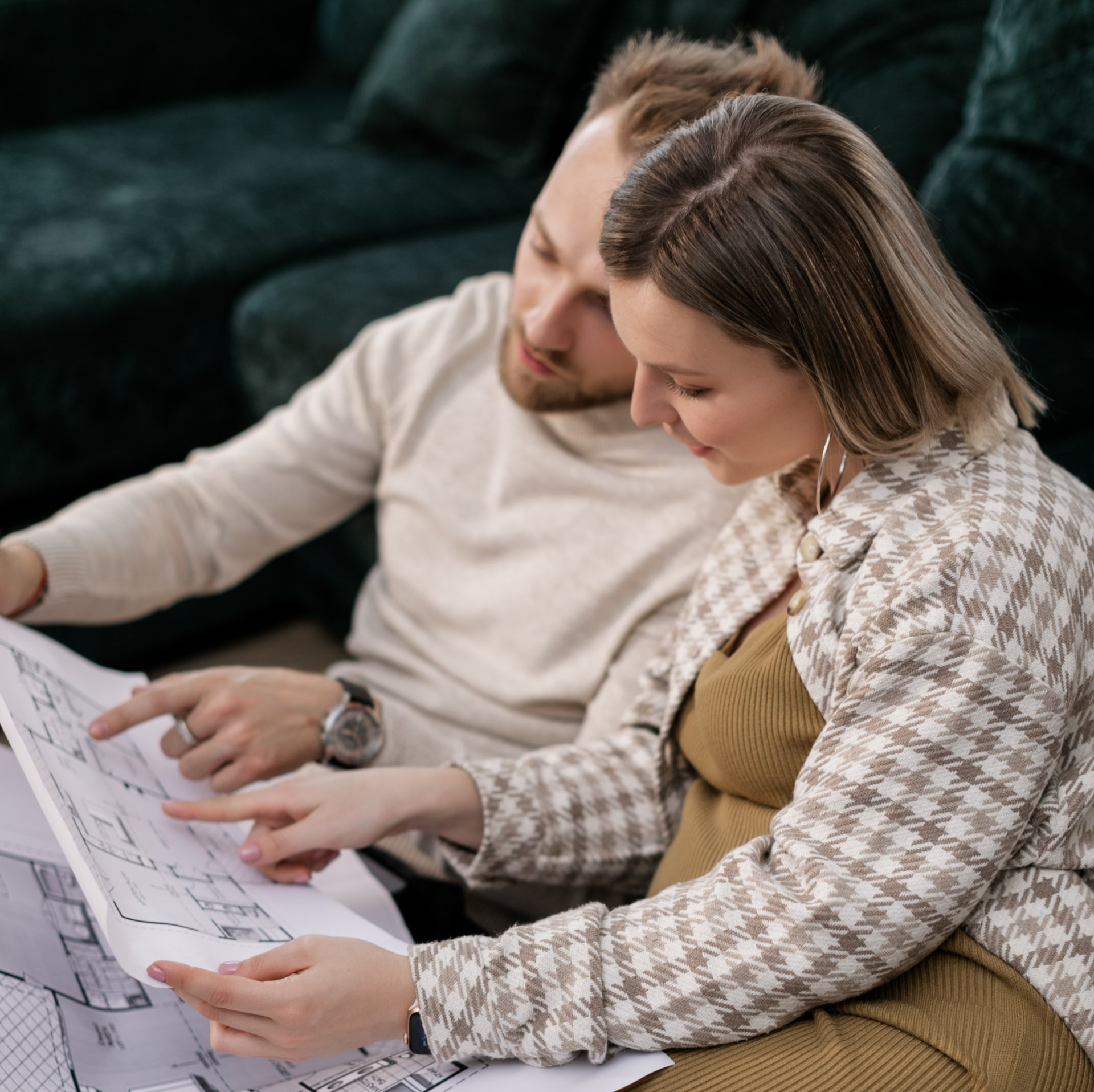 Couple reviewing blueprints, man pointing, woman smiling. Indoors with green sofa.