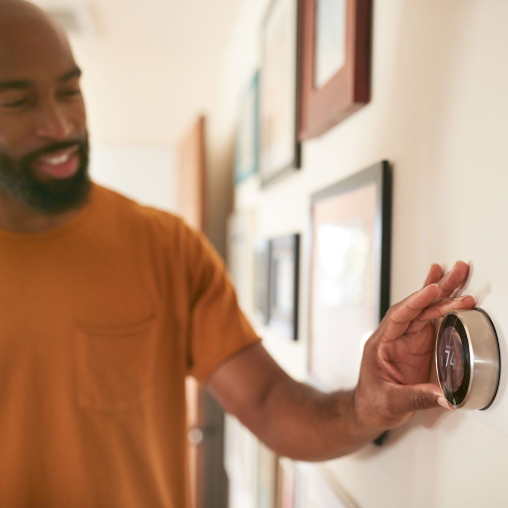 Man adjusting a circular thermostat on a wall, smiling. The setting is indoors with framed artwork.
