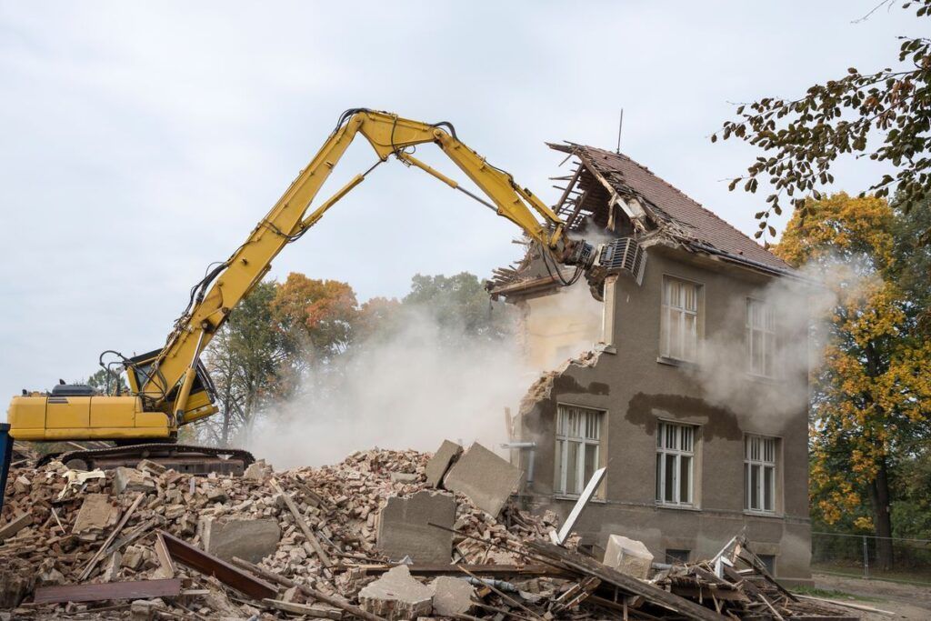 Excavator demolishing a house, with rubble and dust clouding the site.