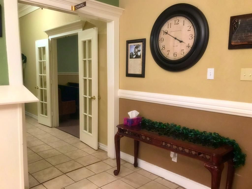 A hallway with tile floors, a wooden console table with a green garland, a wall clock, and a doorway to another room.