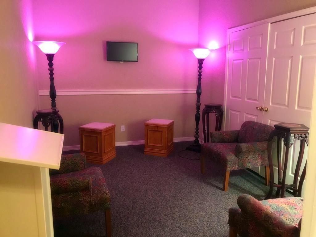 A waiting room with two floor lamps casting pink light, wooden cubes, upholstered chairs, and a television on the wall.
