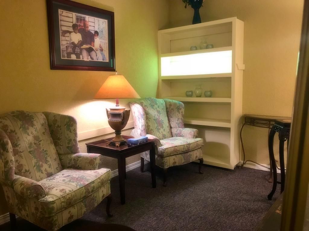 Two floral wingback chairs face each other with a small table and lamp in a room with a white bookshelf and wall art.