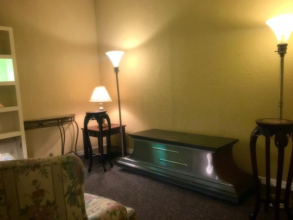 A dimly lit room featuring a floral armchair, a small side table with a lamp, a long bench, and two floor lamps.