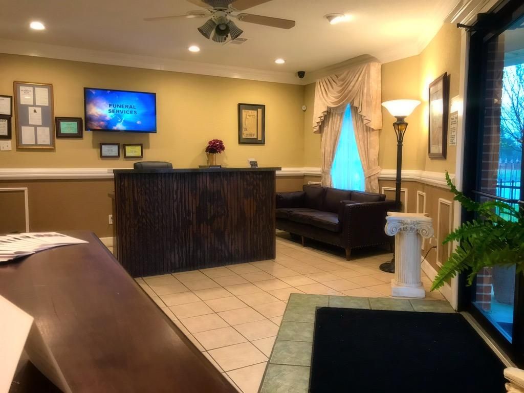A reception area with a dark wood desk, a small couch, a floor lamp, and a TV mounted on a tan wall.