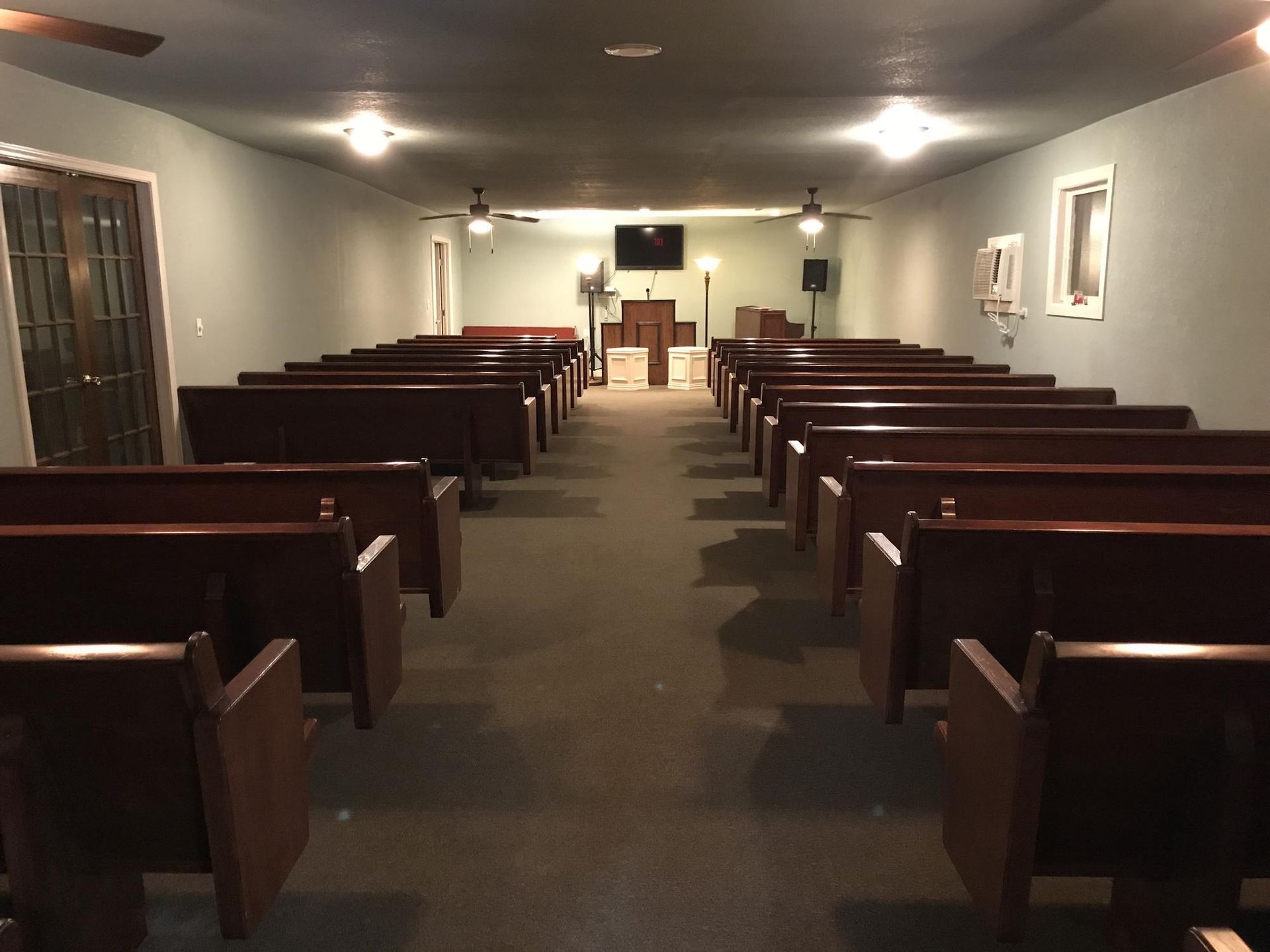Empty church sanctuary with rows of dark wooden pews facing a small altar area with a television screen.