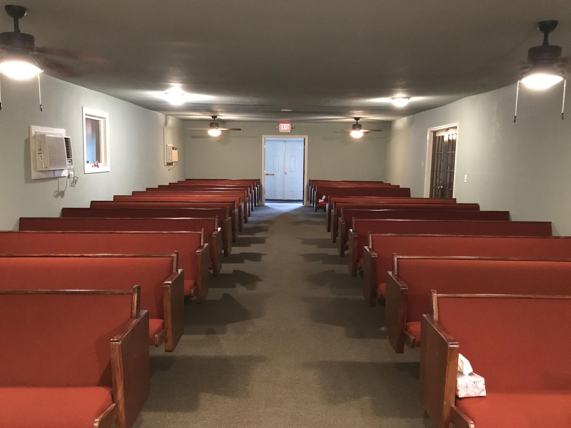 A view down the center aisle of a small church or meeting hall with rows of red padded pews on either side.