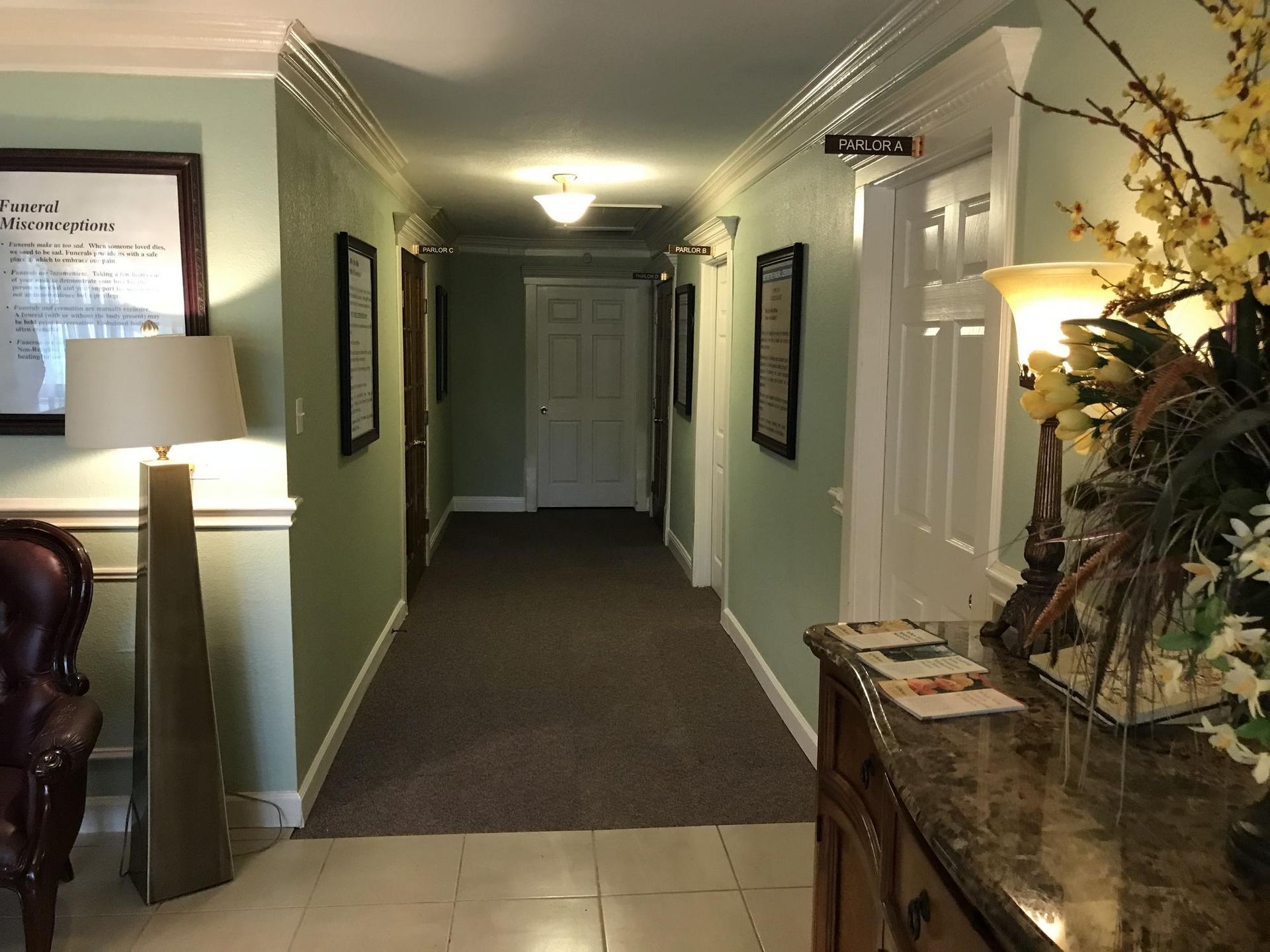 A long hallway with soft green walls, a gray carpet, two lamps, a marble-topped console table, and framed wall decor.