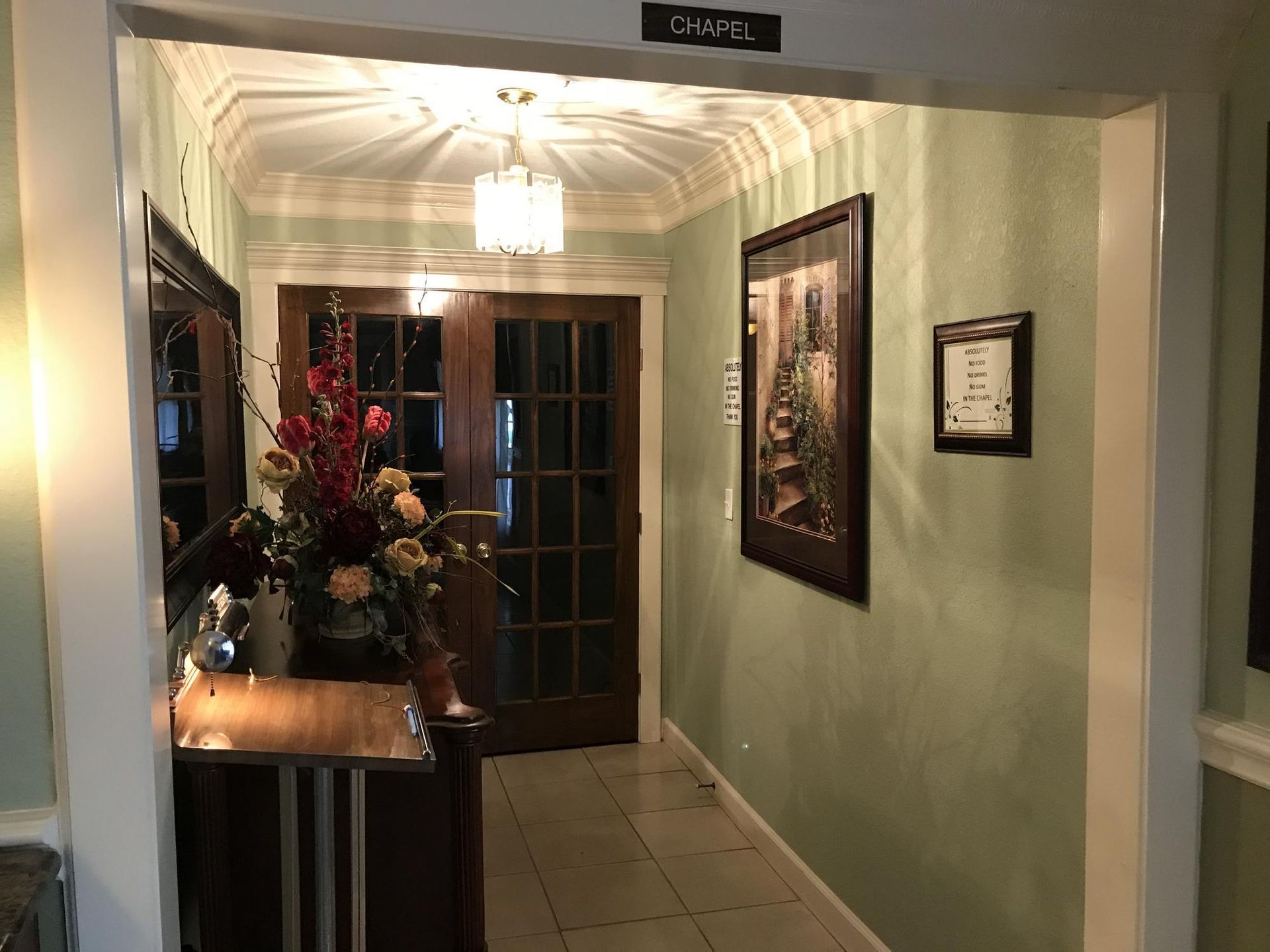 A hallway with light green walls leads to dark wood double doors with glass panes, lit by a crystal pendant ceiling light.
