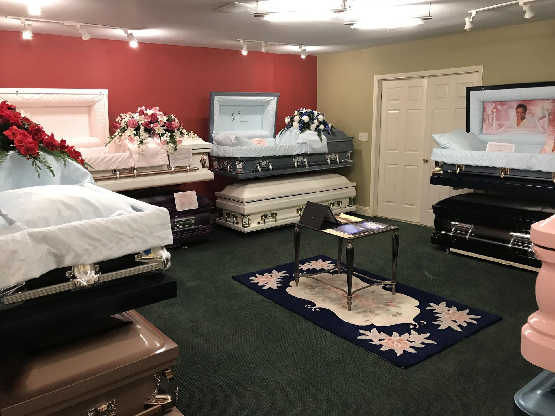 A showroom filled with various open and closed caskets, displayed with floral arrangements on a dark green carpet.