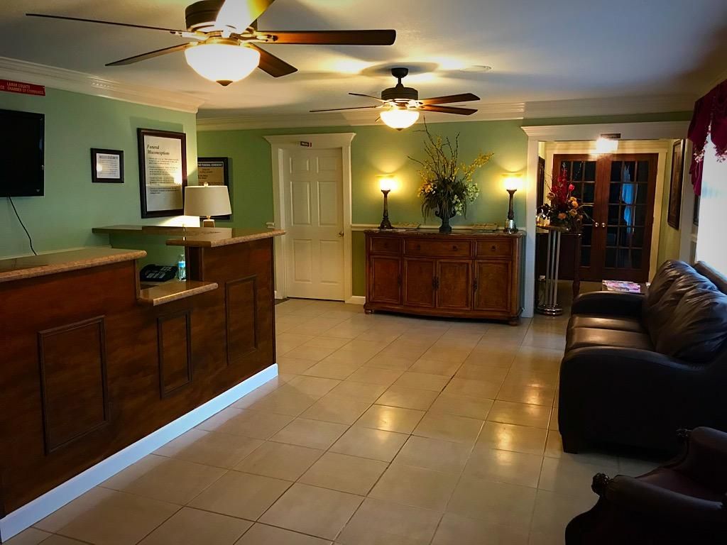 A hotel lobby with a wooden reception desk, light green walls, ceiling fans, a dark leather sofa, and a wooden sideboard.