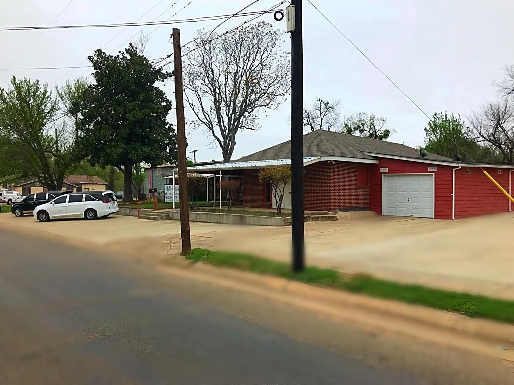 A red brick, single-story commercial building with a white garage door, seen from a street with parked cars on a cloudy day.