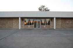 A single-story commercial building with a beige stone exterior, large central windows, and a wide parking lot out front.