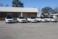 A row of five white funeral limousines parked in front of a tan brick building on a clear, sunny day.