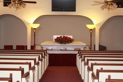A casket topped with red roses sits in front of a church altar between two floor lamps, with pews facing it.