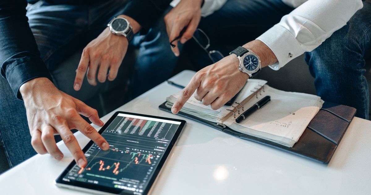 Two people point at a tablet displaying stock charts while sitting at a table with a notebook. Both wear watches.