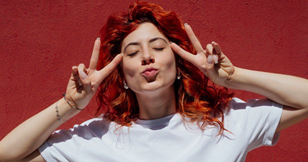 Woman with red hair making a peace sign, blowing a kiss. Against a red wall.