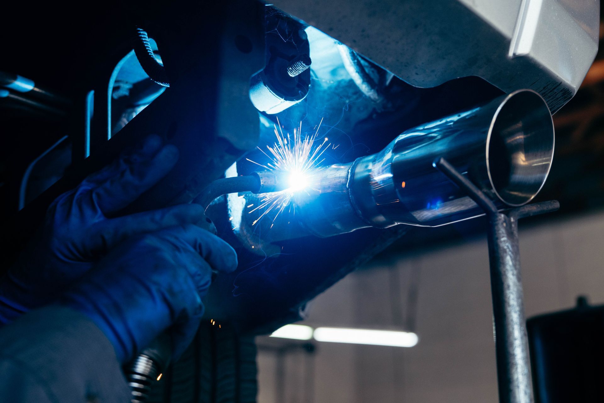 A worker in blue gloves uses a welding torch to attach a metal exhaust pipe to the underside of a vehicle.