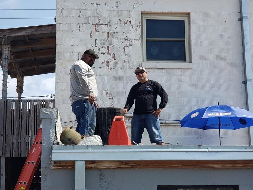 Two workers in casual clothing stand on a flat roof next to a blue umbrella and a red container against a white wall. Two workers in casual clothing stand on a flat roof next to a blue umbrella and a red container against a white wall.