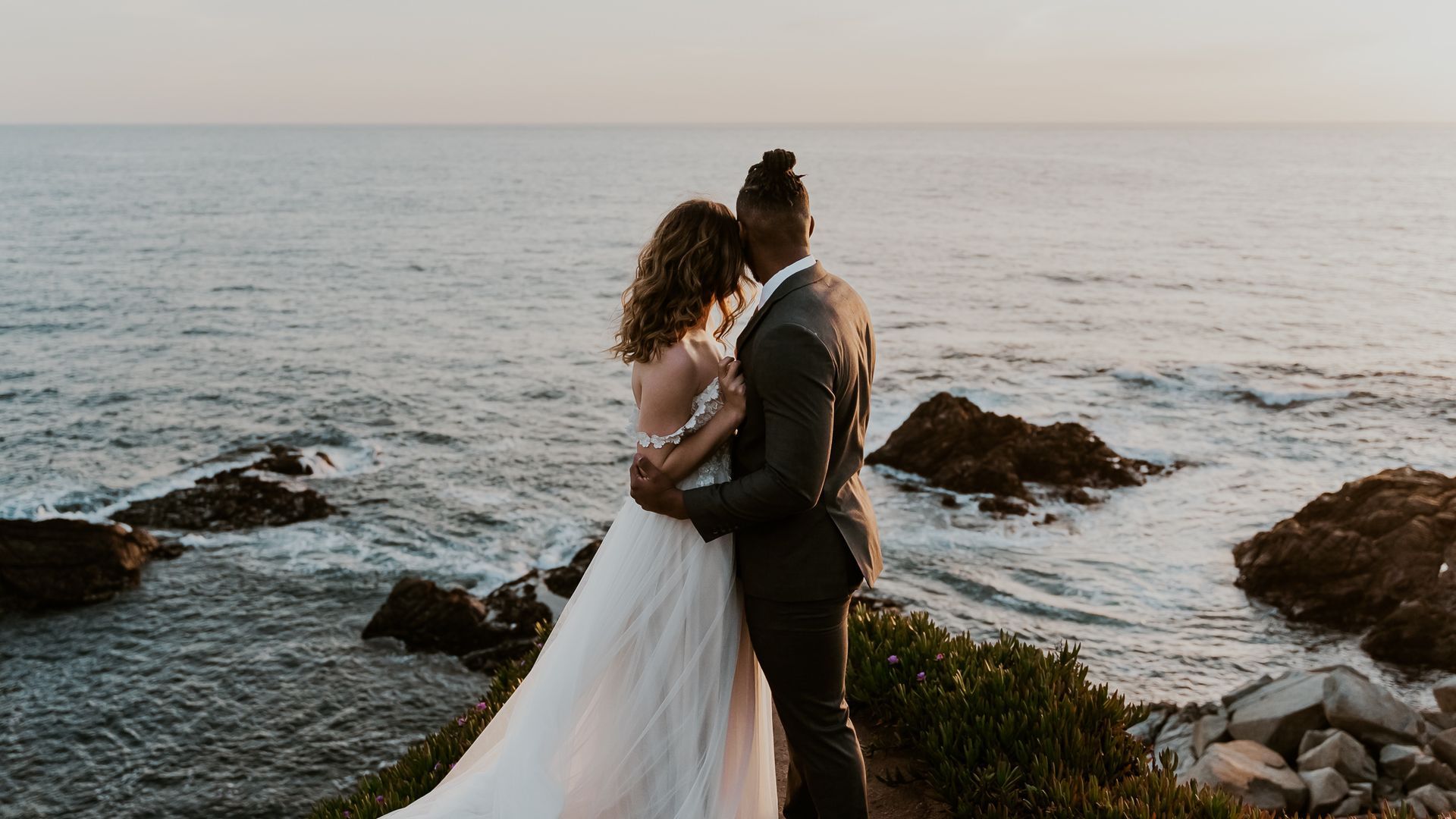 A bride and groom are standing on a rocky cliff overlooking the ocean.