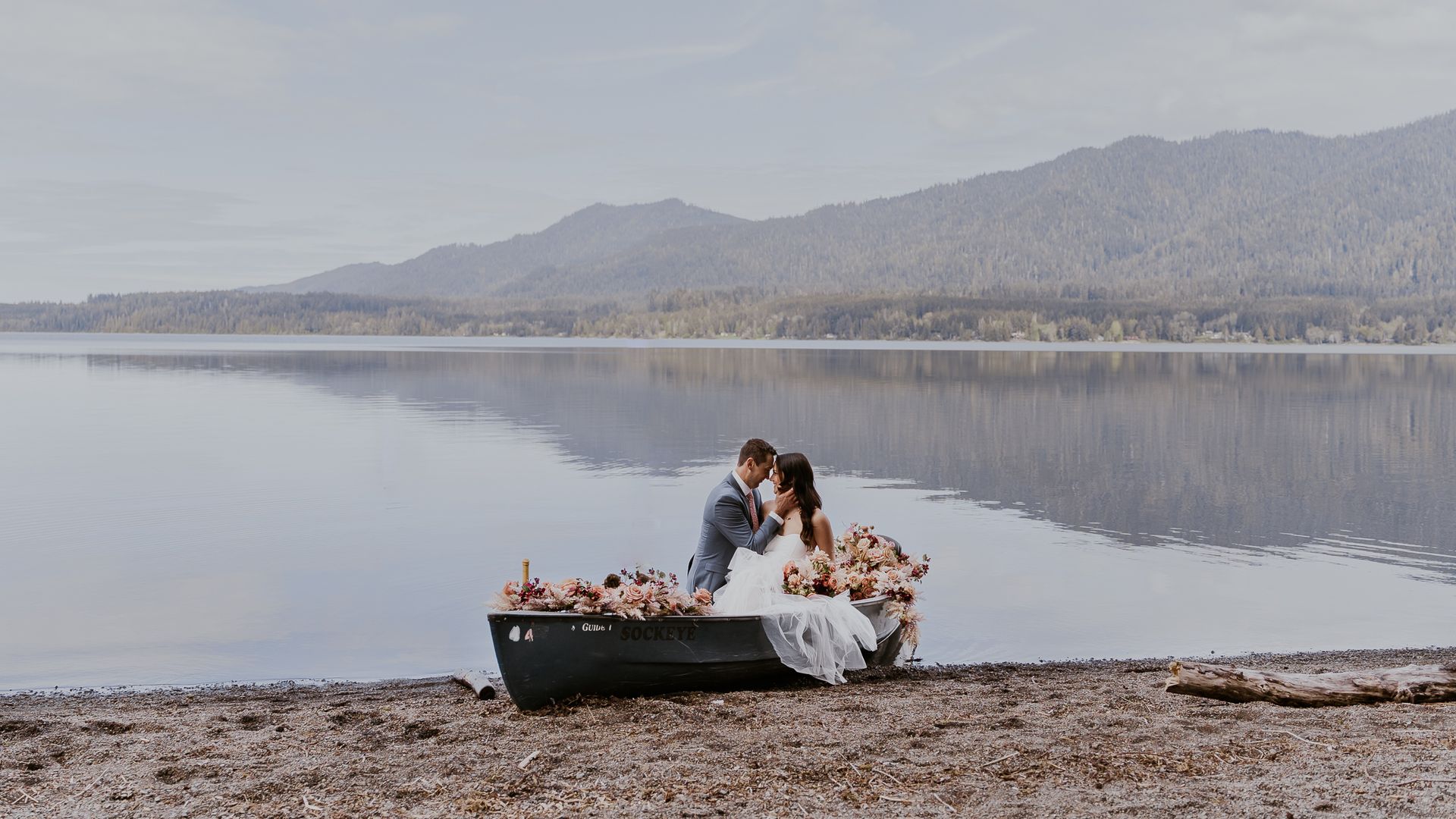 A bride and groom are sitting in a boat on the shore of a lake.