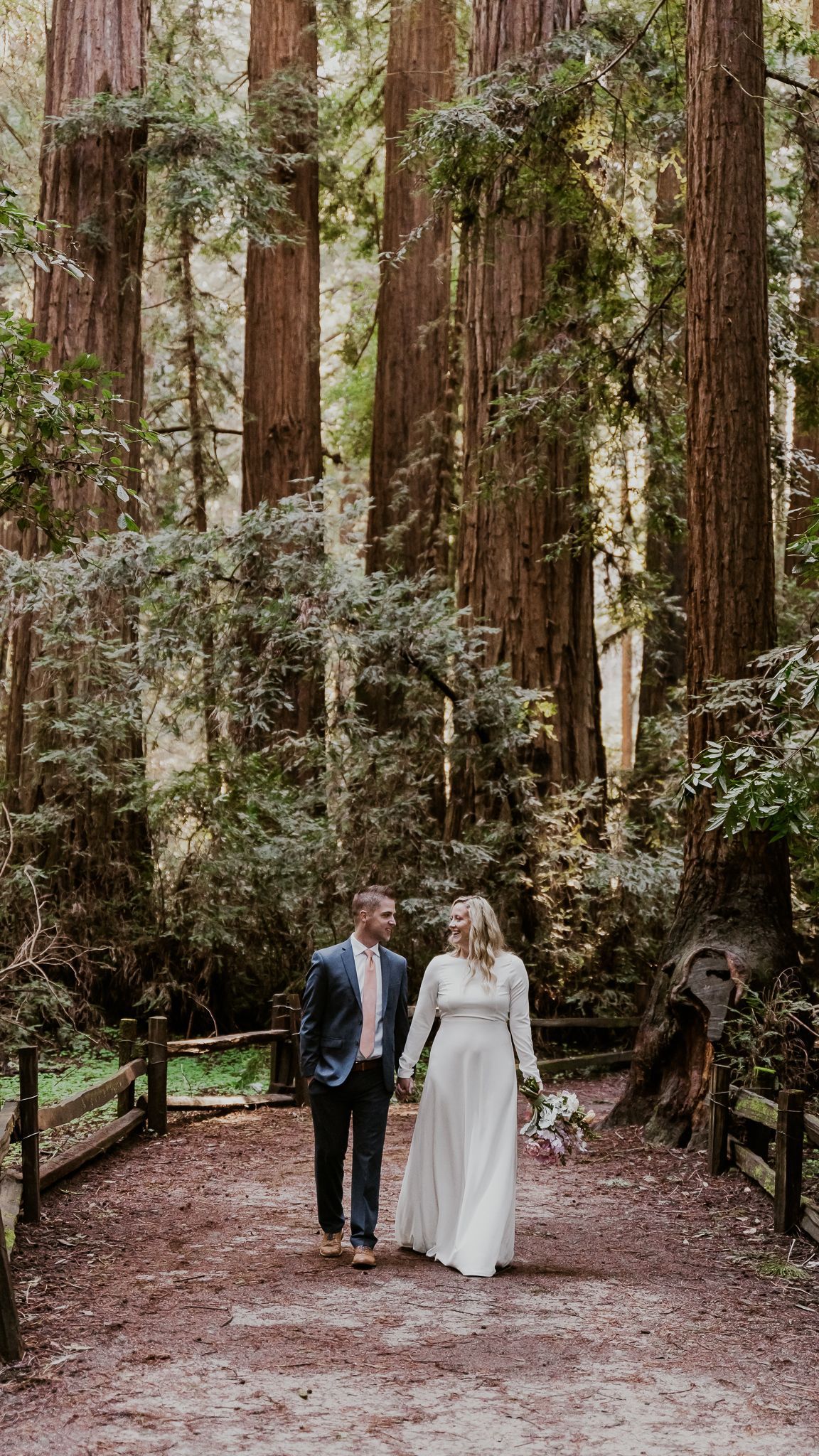 A bride and groom are walking through a forest holding hands.