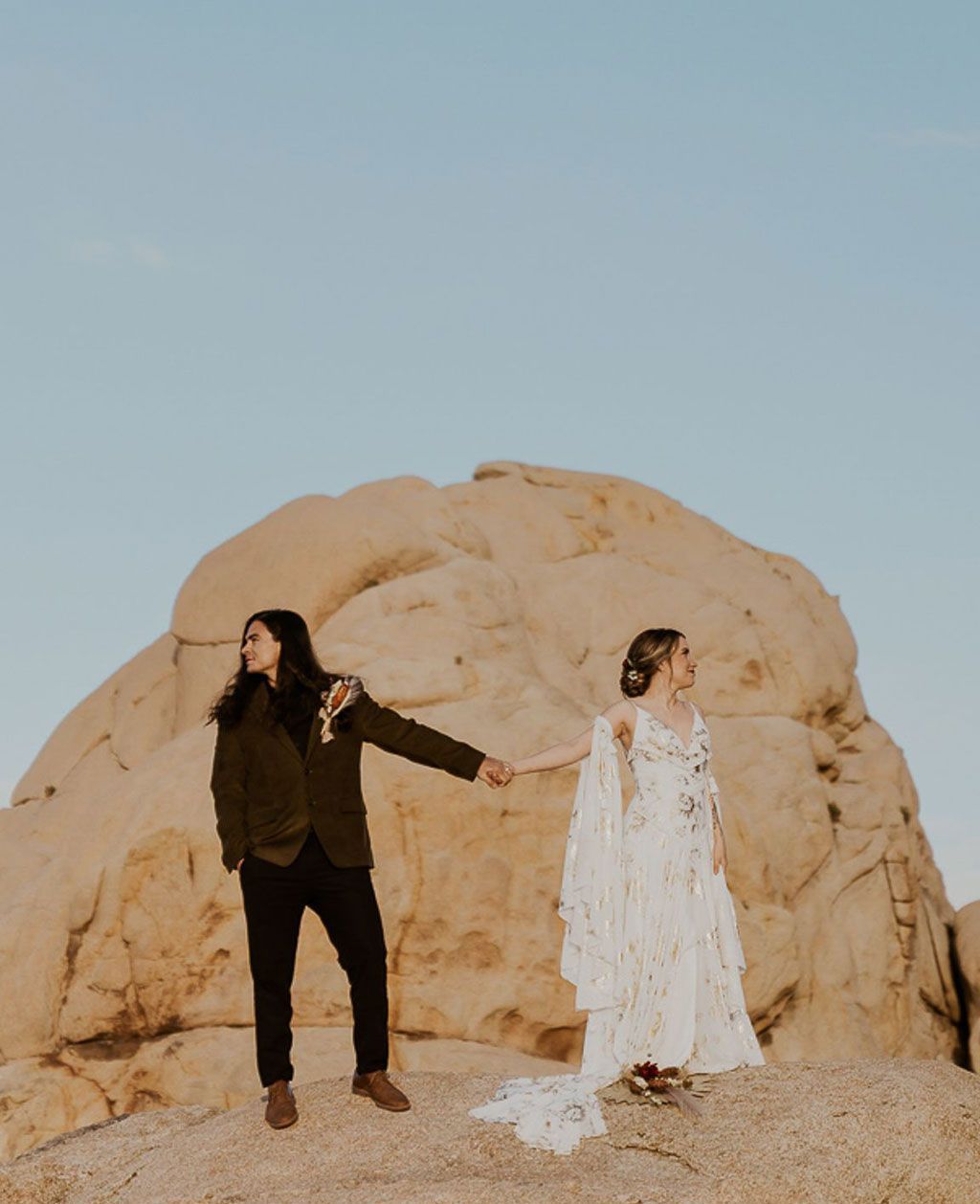 A bride and groom are holding hands in front of a large rock.