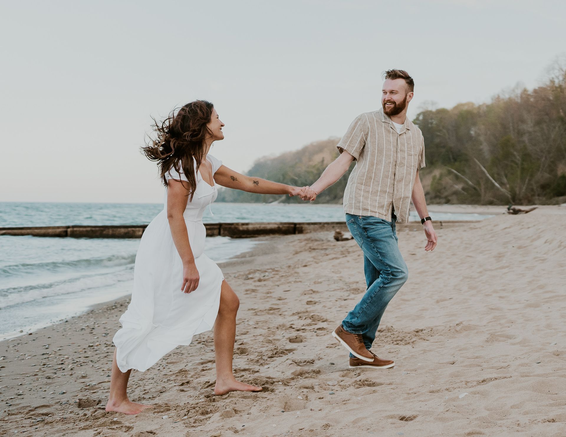 Couple holding hands, walking on a beach. The woman is wearing a white dress, the man jeans and a shirt.