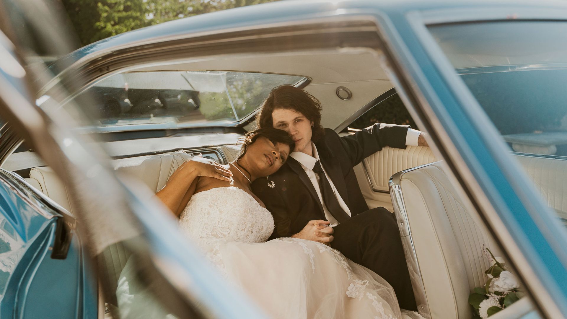 A bride and groom are sitting in the back seat of a blue car.